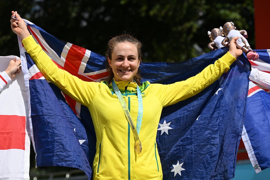 An Australian female road cyclists stands with the national flag behind her while wearing her Comm Games gold medal.