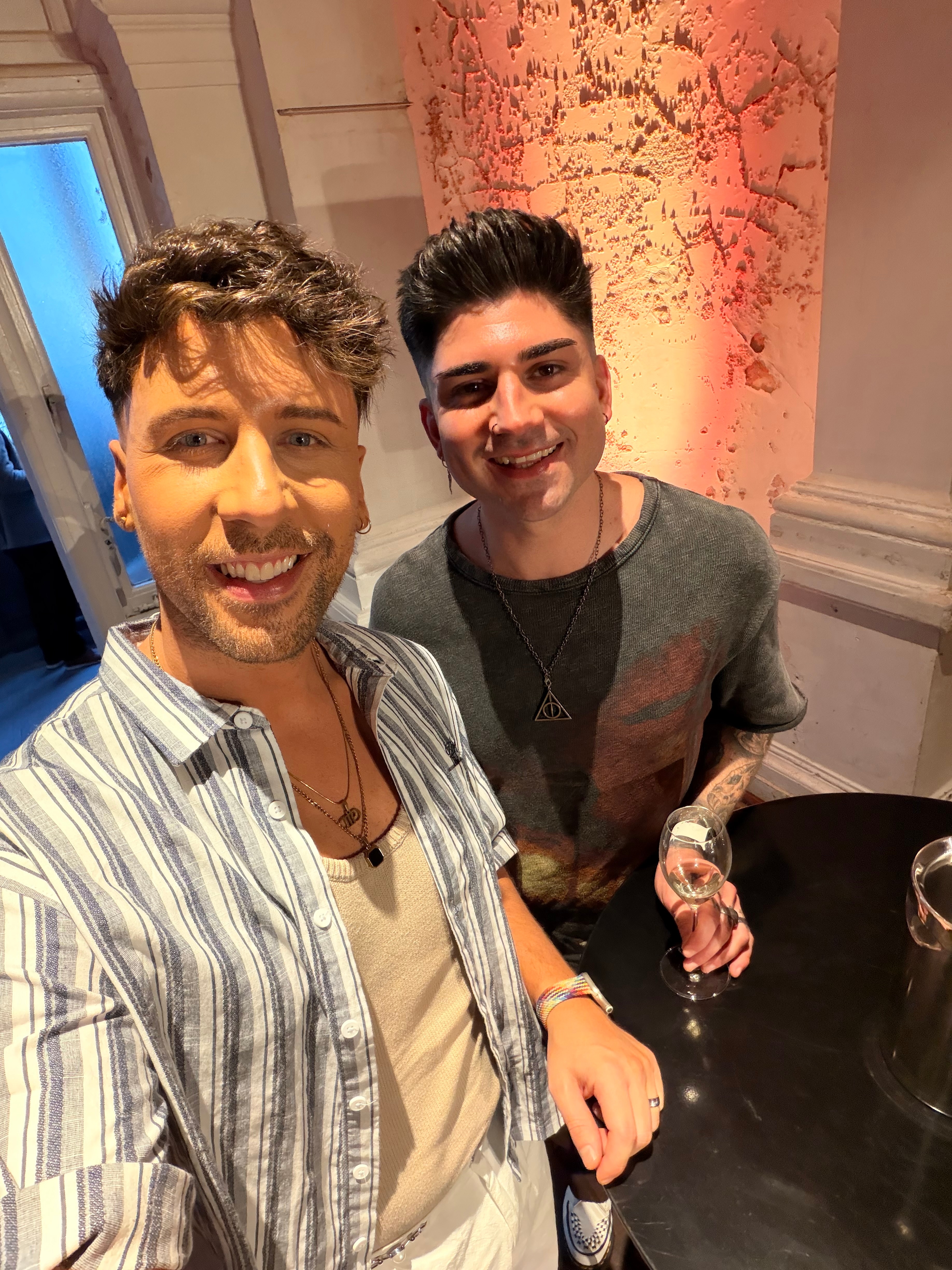 Dos jóvenes sonrientes, ambos con el pelo corto y oscuro, tomándose un selfie en el interior.