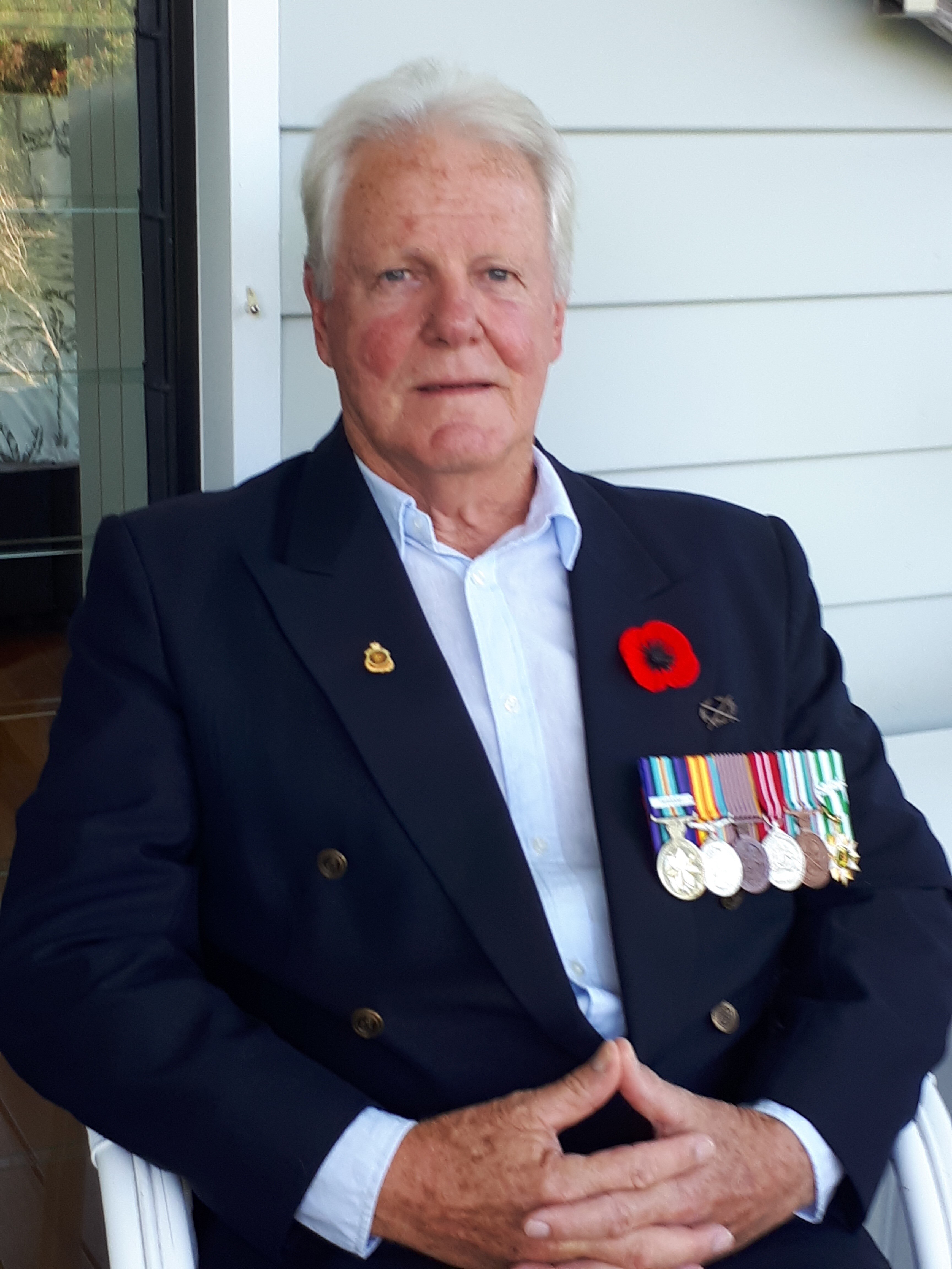 A man sitting in a chair wearing a navy blazer with a poppy and war medals pinned on his chest.