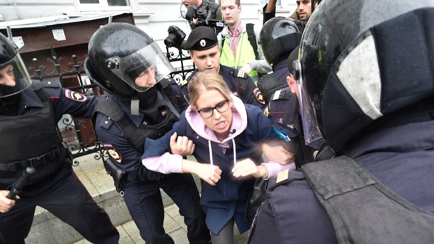 Police officers with helmets and batons detain an opposition candidate as she struggles against their grip.