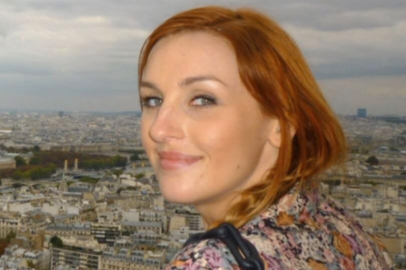 A tight head shot of a smiling young woman with red hair posing for a photo with a city behind her.