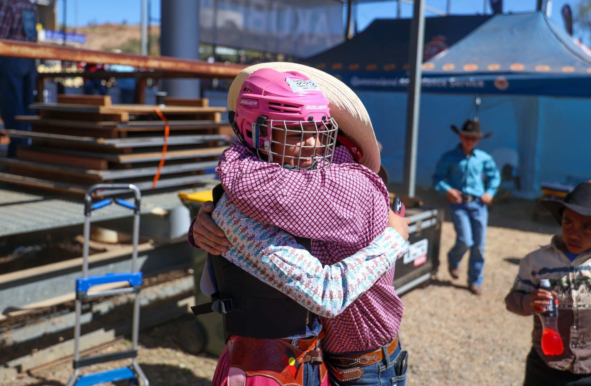 A young rider wearing a pink helmet and protective vest hugs another person beside the rodeo arena.