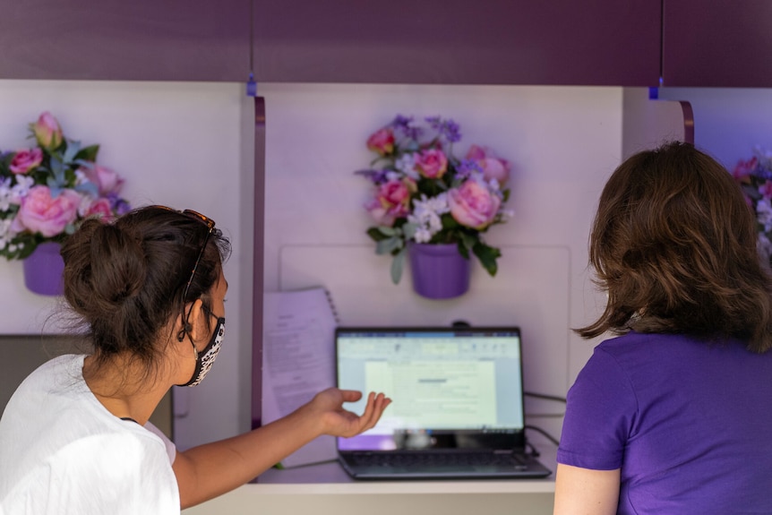 The back of two women's heads looking at a computer screen with purple flowers on the wall nearby.