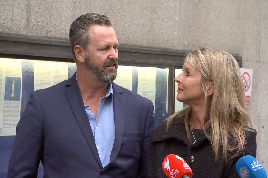 A middle-aged couple stand in front of a concrete building.