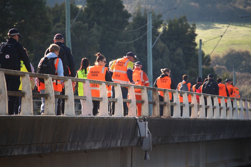 Back view of a group of people in orange and yellow search vests and two police officers walk across a bridge.