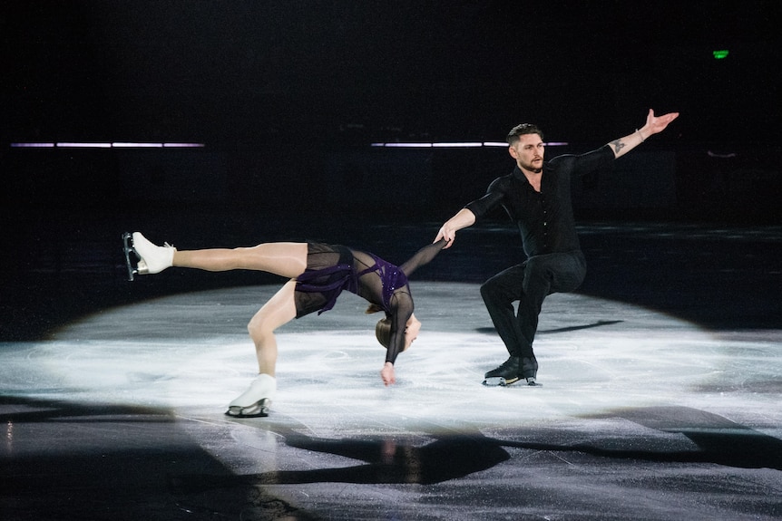 Harley Windsor on a spotlight-lit ice rink, his right hand holds on to his skating partner's hand as she backbends.