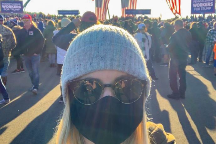 A photo of a young woman in sunglasses, a beanie and a face mask with US flags and crowds behind her