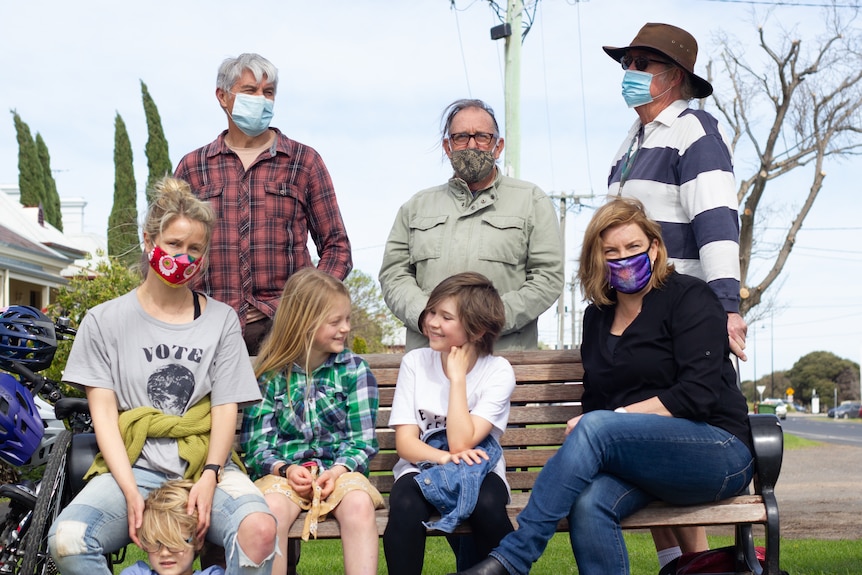 Two women three men and two children sit and stand together outside with face masks on. 