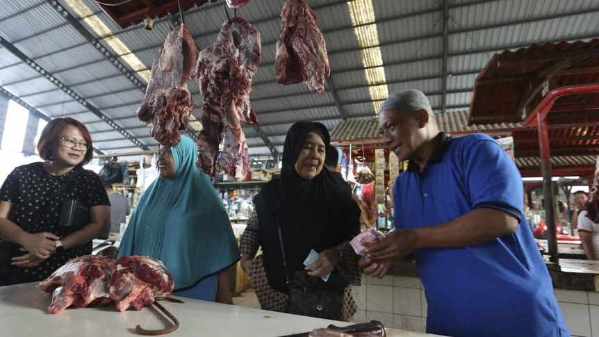 Three women and a butcher exchange cash at a market stall lined with meat on hooks at an undercover market.
