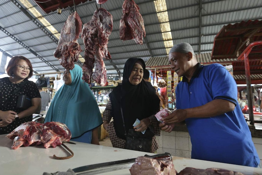 Three women and a butcher exchange cash at a market stall lined with meat on hooks at an undercover market.