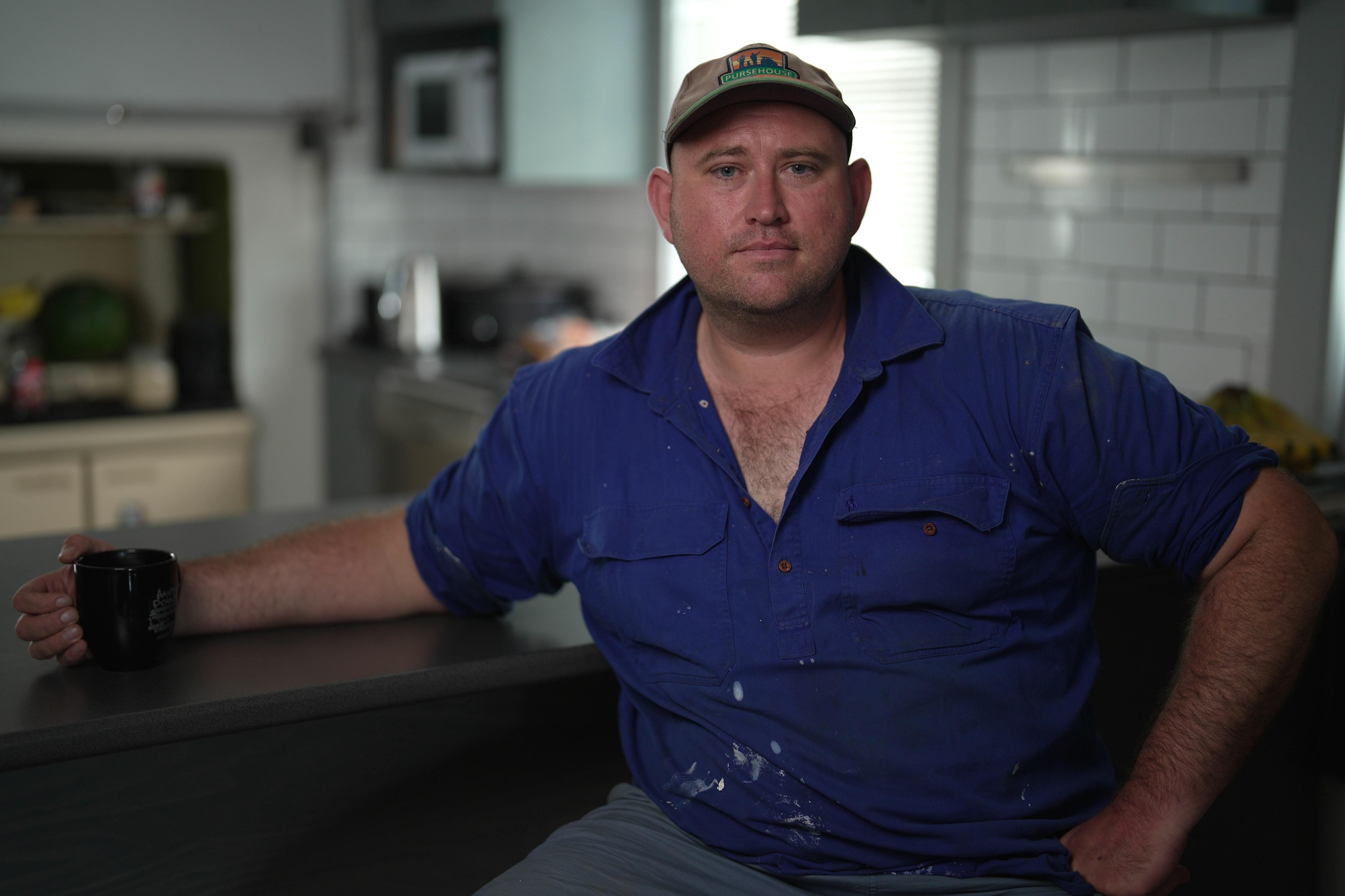 Rohan, wearing a blue work shirt and cap, sits at his kitchen table with a mug.