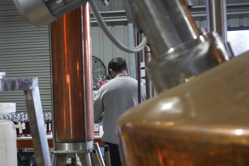 a man with his back to the camera works on a bench surrounded by distillery equipment 