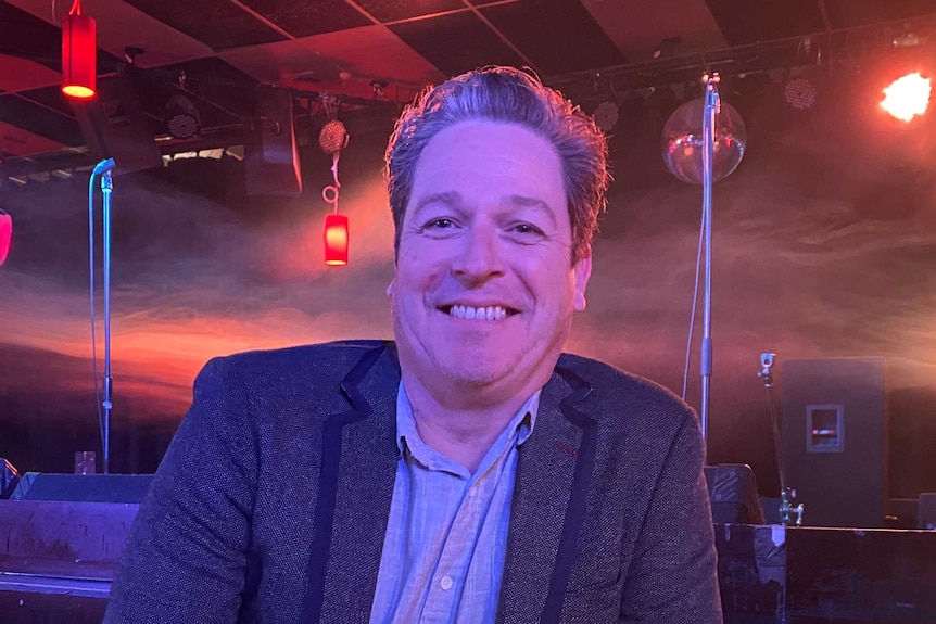 A smiling man wearing a jacket and open necked shirt smiles in a club.