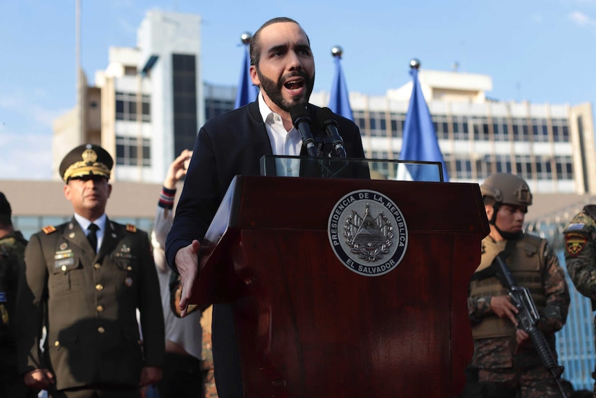 El Salvador's president wears a black suit speaks while standing at a lectern.