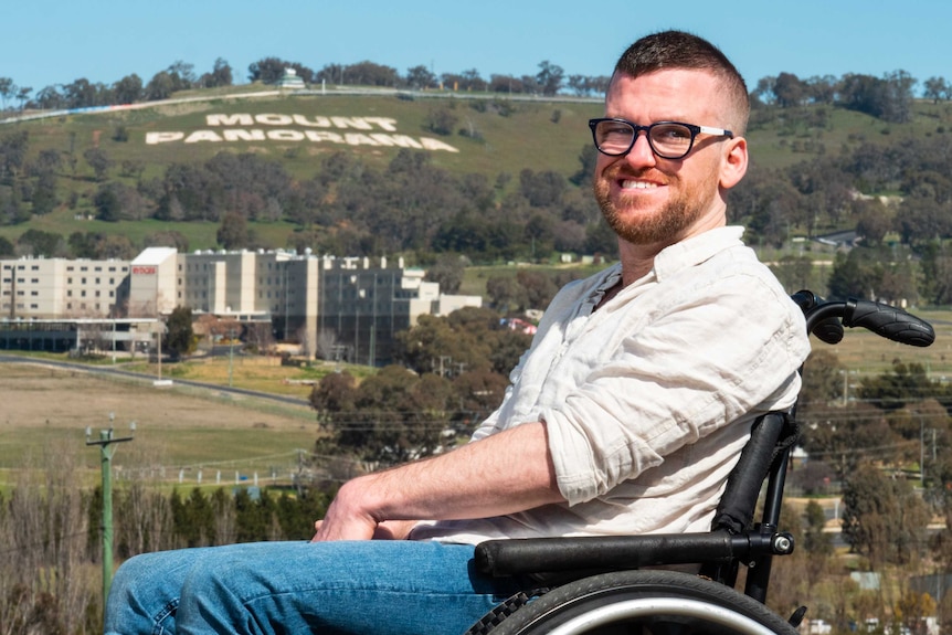 A man sits side on to the camera with Mount Panorama in the background.