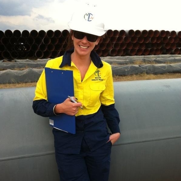 A woman in a hard hat, sunglasses and bright yellow collared shirt leans against a large pipe holding a clipboard.