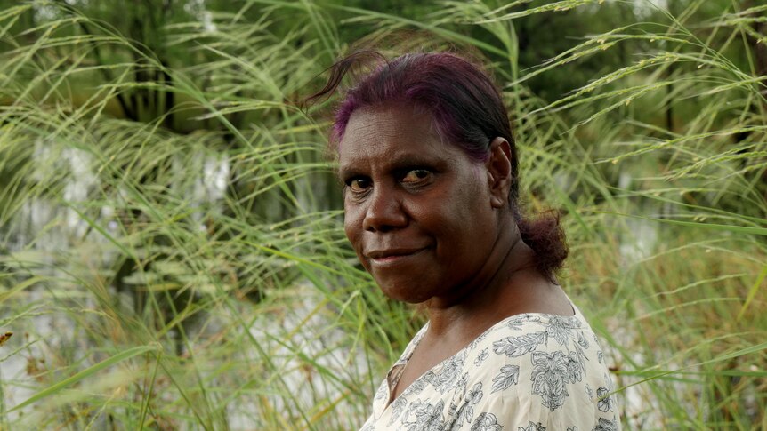 A woman with purple hair sits with a lush green billabong in the background. 