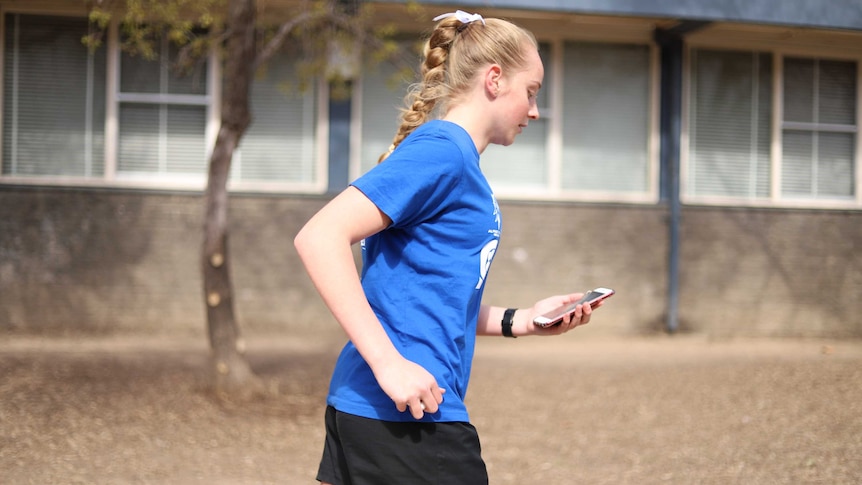 Blonde-haired girl with blue dress jogs past school building while holding and looking at mobile phone.