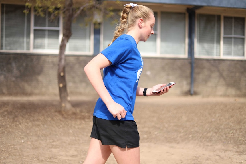 Blonde-haired girl with blue dress jogs past school building while holding and looking at mobile phone.