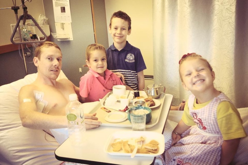 A man in a hospital bed with a meal tray on a table, with three children around him.