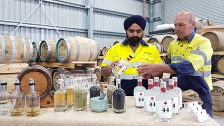 Photo of two men in a large shed with barrels and bottles of liquor around them. One bald older man, one bearded man in turban