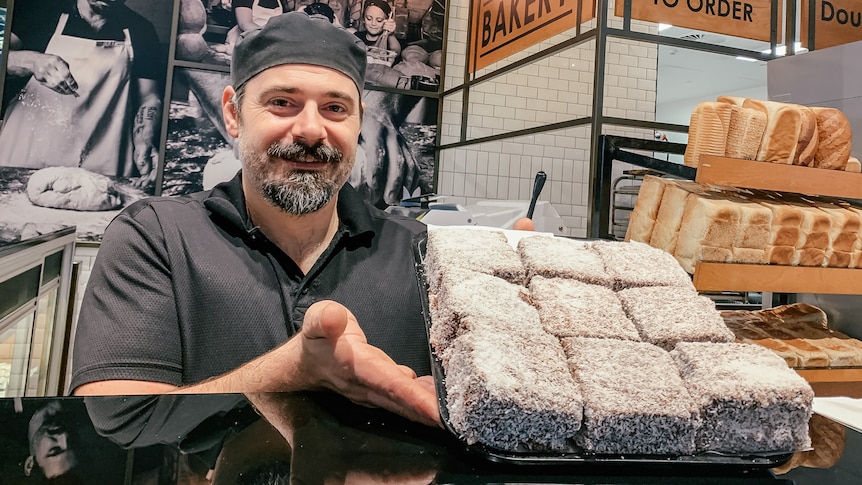 A man wearing black baker's, tshirt, holds tray of lamingtons at the counter of a bakery, breads on shelf, posters on wall.