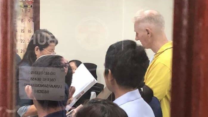 A photo taken through a window shows Peter Scully standing in a court room with a woman appearing to read something to him.