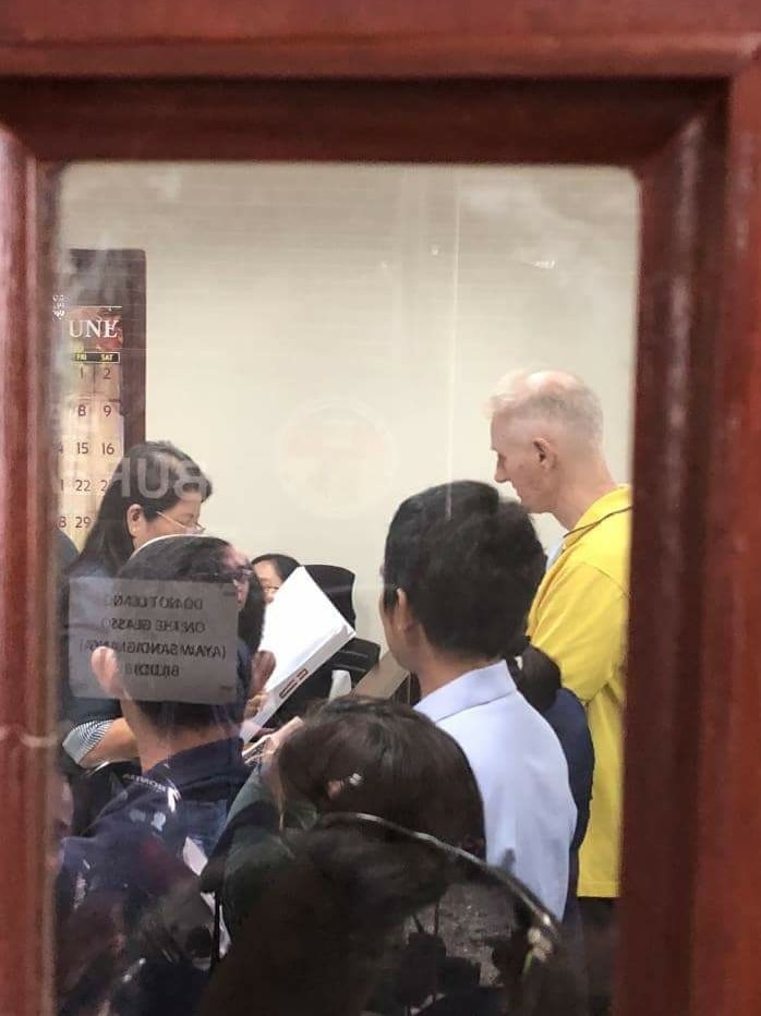 A photo taken through a window shows Peter Scully standing in a court room with a woman appearing to read something to him.