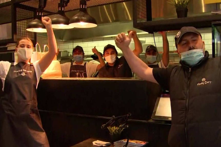 Five workers wearing aprons with the words "Sharing the love" do the thumbs up and sheer in a restaurant kitchen.