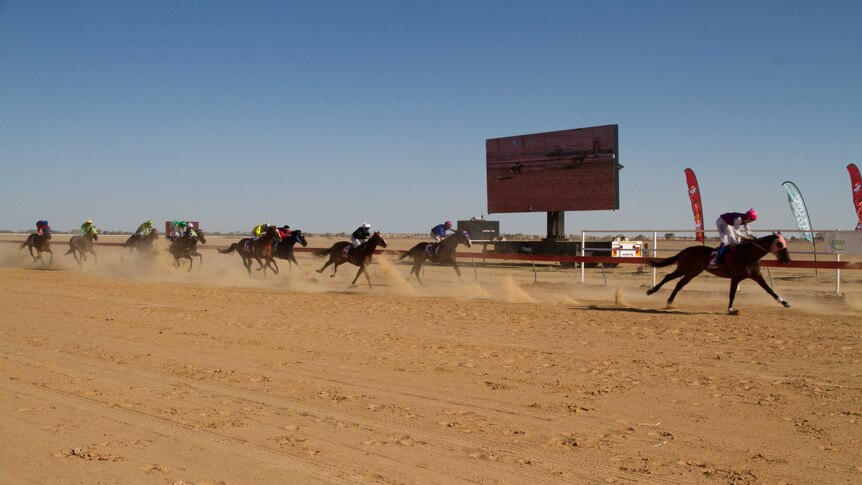Horses and jockeys race along a red dirt track kicking up dust.