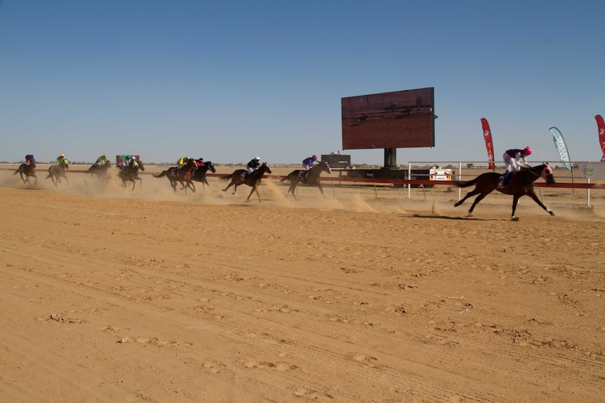 Horses and jockeys race along a red dirt track kicking up dust.