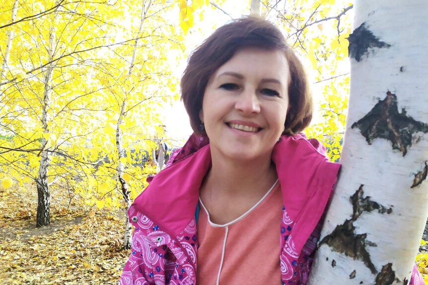 A photo of a woman smiling with short hair with a tree and yellow leaves in the background.