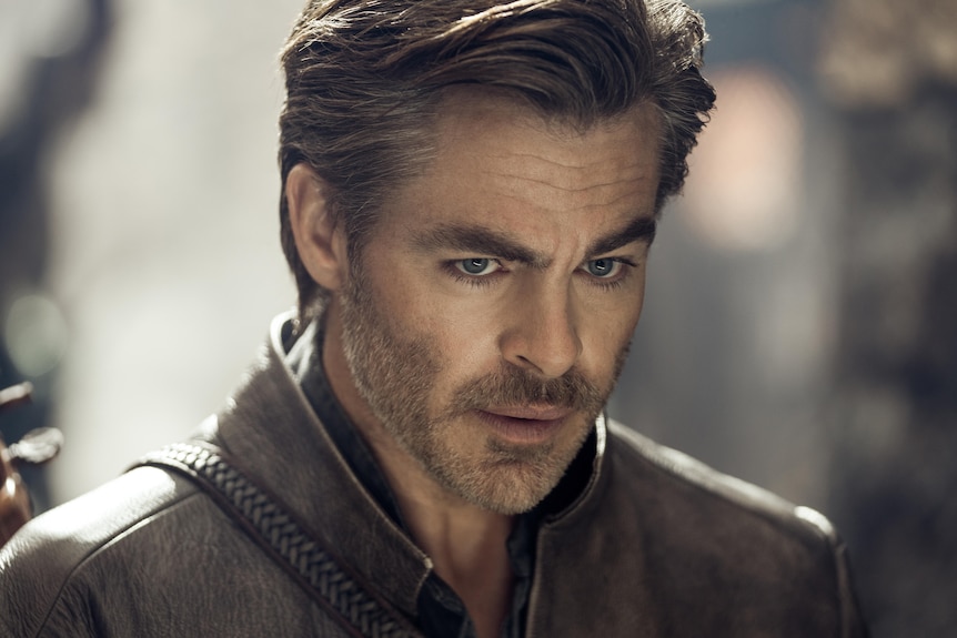 A close up of a white middle-aged man with blue eyes, greying hair and stubble wearing a brown leather vest.