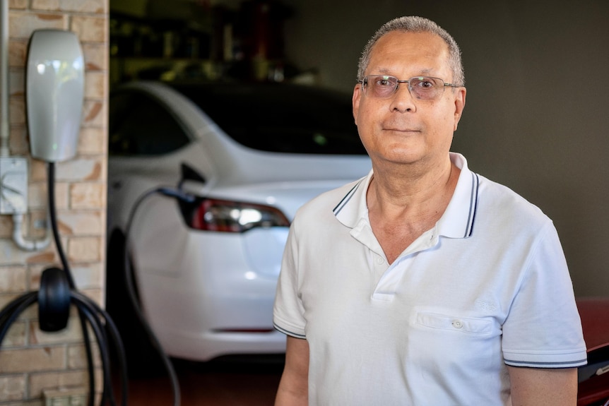 a man wearing a white polo shirt standing in the doorway of a garage. a white tesla can be seen in the background