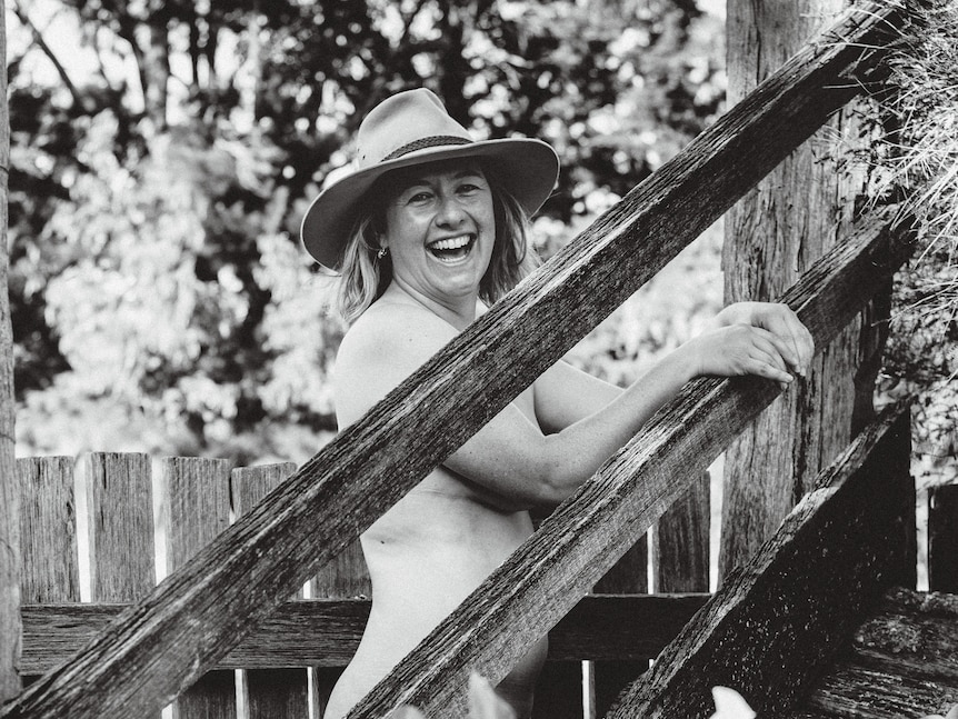 A black and white image of a naked woman, standing behind a wooden fence. 