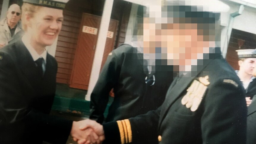 A young woman in a Navy uniform smiles and shakes the hand of an officer, whose face is pixellated.