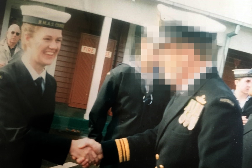 A young woman in a Navy uniform smiles and shakes the hand of an officer, whose face is pixellated.