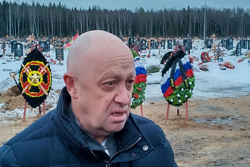 A bald, white man stands in front of wreaths of flowers on a snowy landscape.