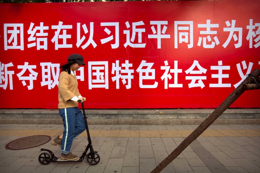 A woman is seen riding a scooter past a banner written in Chinese charachters.