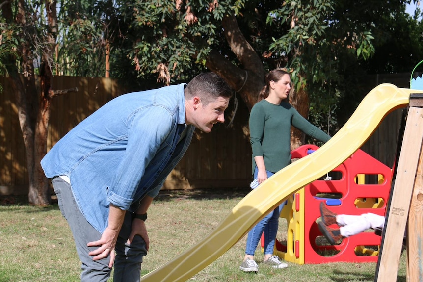 A dad watches his child play near a yellow slide at play equipment.