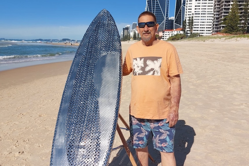 An image of Eddie Rigo standing on the beach with a metal surfboard 