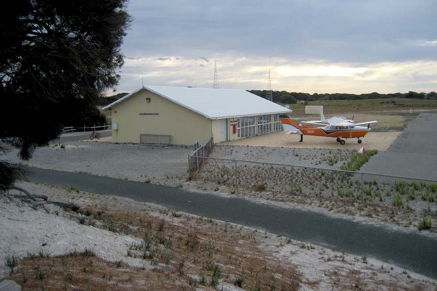 A light plane sits in front of a small brick building next to a short airstrip.