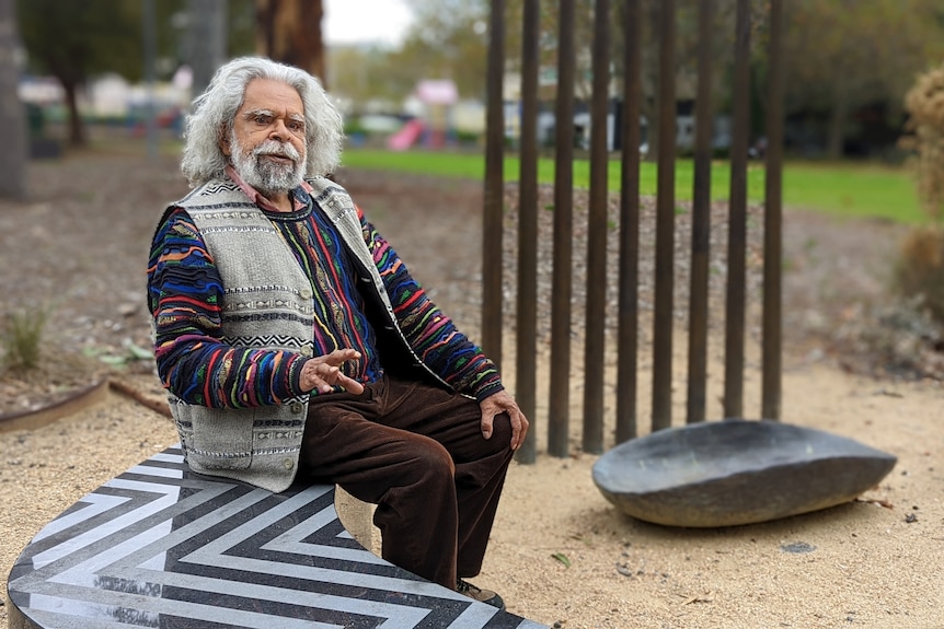 An older man sits in a park, speaking to someone behind the camera. 