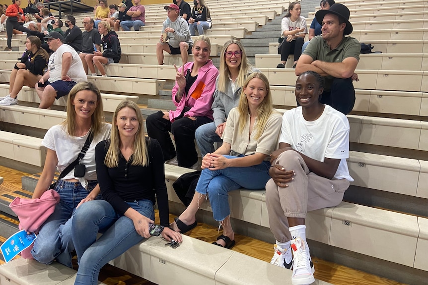 Six women and a man sit together in the bleachers at a basketball game