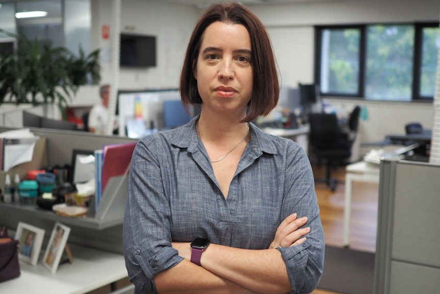 Woman with dark mid length hair, wearing grey shirt, with sleeves rolled up, unsmiling, arms crossed and standing in open office