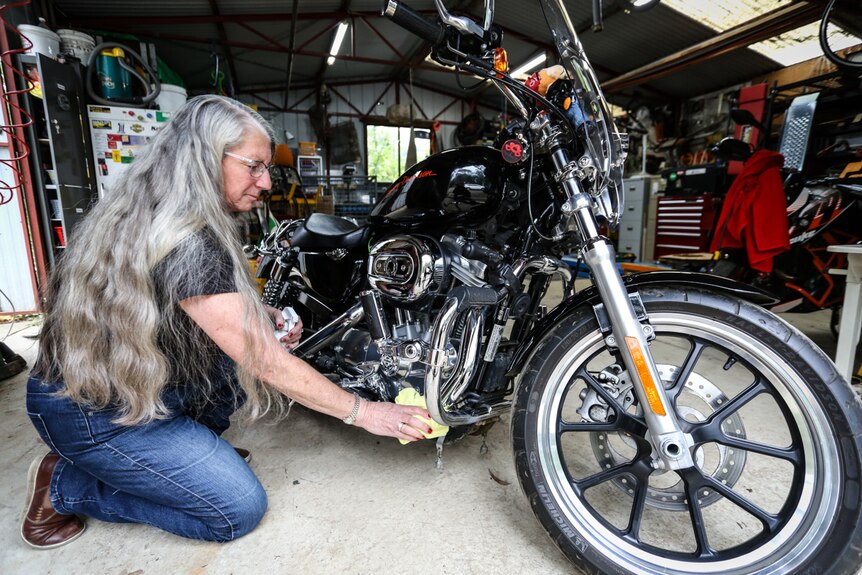 An older woman with very long grey hair shines the chrome on a mean-looking Harley-Davidson.