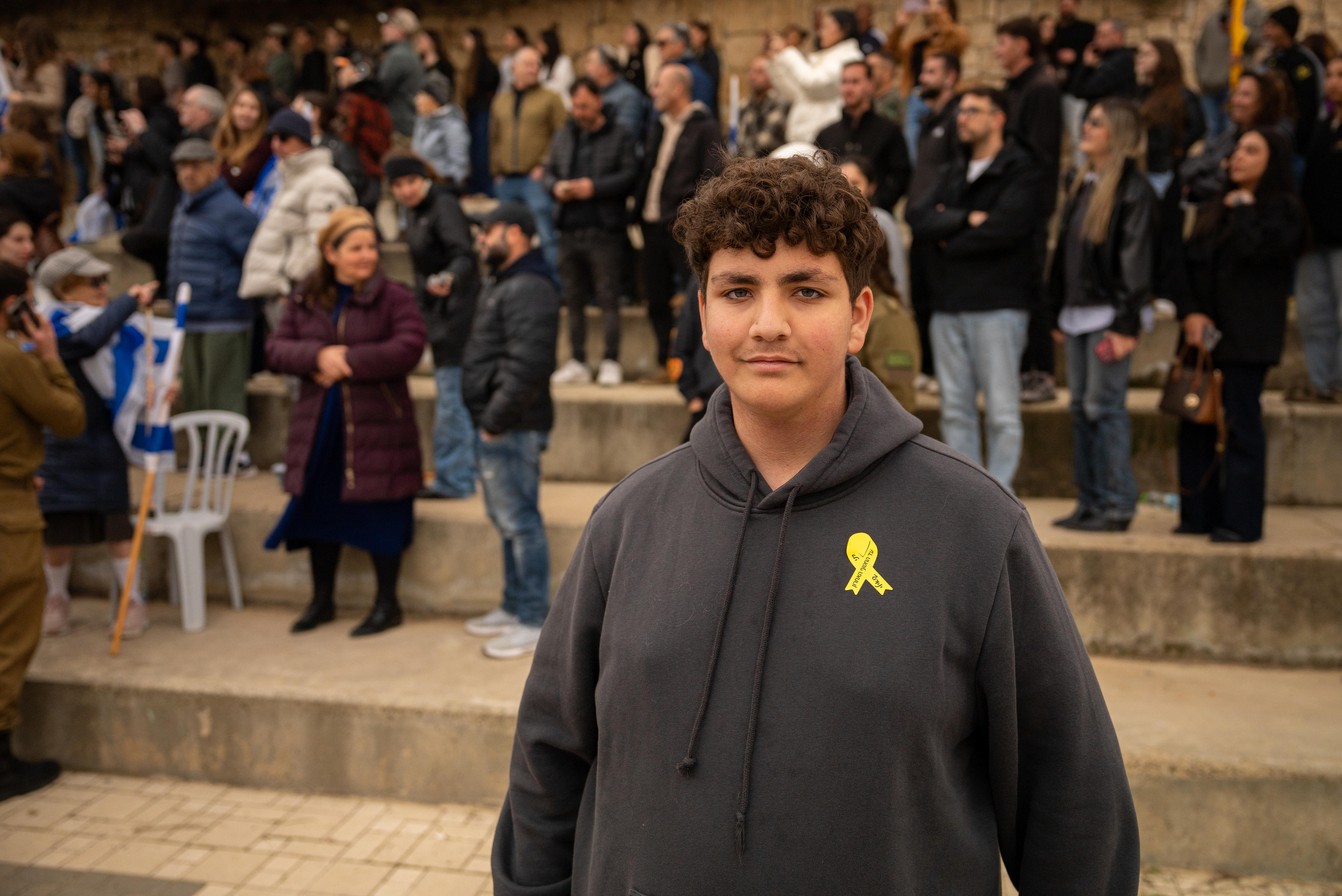 Um adolescente com cabelos castanhos cacheados, sobrancelhas, vestindo um suéter preto e um alfinete de fita amarelo.