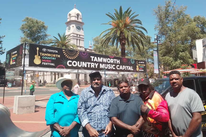 Five band members standing outside of Tamworth Country Music Festival with a banner in the background.
