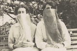 Two sisters sit on a park bench wearing long masks during the flu pandemic in 1918.