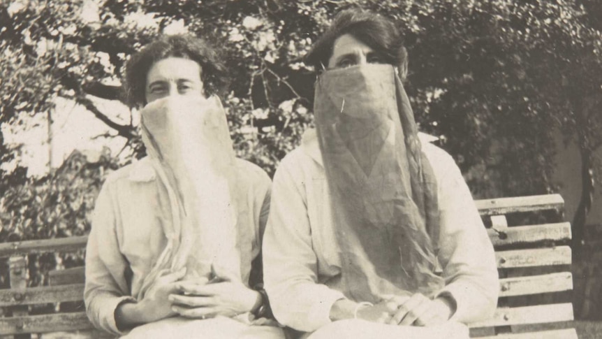 Two sisters sit on a park bench wearing long masks during the flu pandemic in 1918.